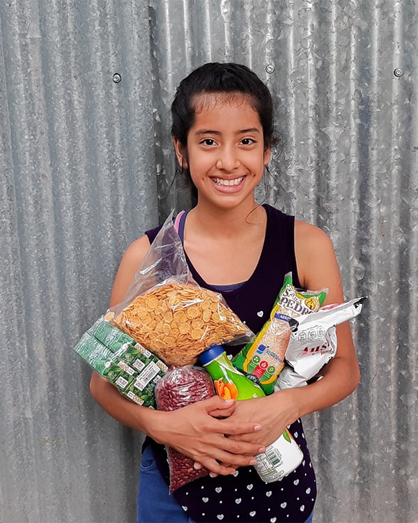Smiling girl in front of a corrugated steel wall holding an armful of food.
